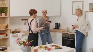 Kırmızı şarap içip sohbet eden, modern minimalist mutfağın önünde duran üç güzel kadının orta boy fotoğrafı.