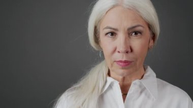 Koyu gri arka planda kameraya gülümseyen uzun gri saçlı çekici olgun bir kadının orta boy yakın portresi.