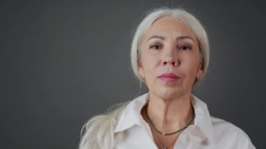 Beyaz gömleğindeki uzun gri yakalı ve koyu gri arka plandaki kameraya gülümseyen kendine güvenen yetişkin bir kadının orta boy yakın portresi.