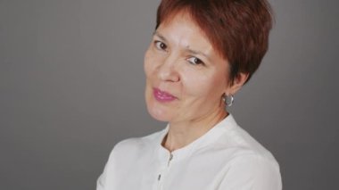 Gri arkaplanda kameraya gülümseyen kısa saçlı olgun kadının orta boy yakın stüdyo portresi.