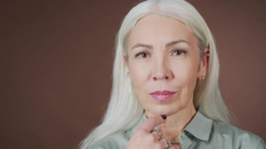 Çekici beyaz kadın portresi. Uzun gri saçlı, kahverengi stüdyo arka planındaki kameraya bakıyor.