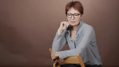 Kameraya poz veren, kahverengi arka planda oturan olgun kısa saçlı kadının orta boy stüdyo portresi.
