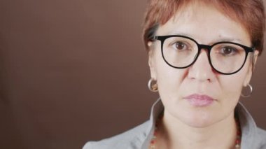 Gözlüklü, kahverengi stüdyo arka planında kameraya bakan üzgün olgun kadının PAN yakın plan portresi.
