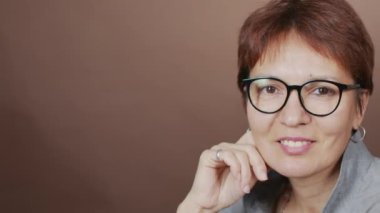PAN, kahverengi stüdyo arka planında kameraya gülümseyen, kendine güvenen gözlüklü yetişkin kadının portresi.