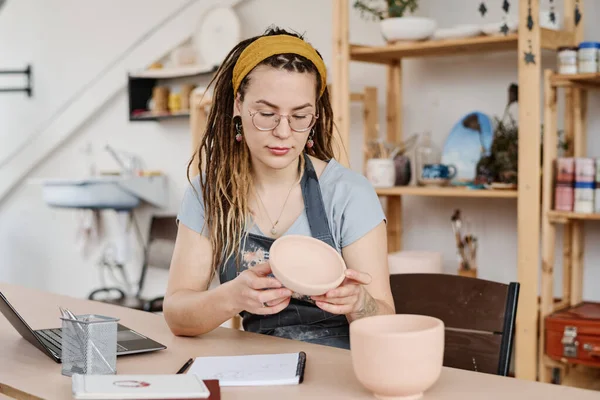工房や工房で土鉢の品質を確認しながら テーブルの上に手作りの土器を持つ若い女性 — ストック写真