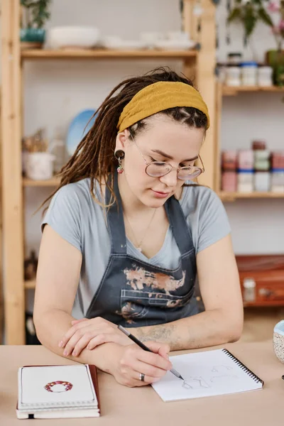 小さな店やスタジオでテーブルに座っている間にノートパッドで陶器製品の若い創造的な女性の陶芸家のスケッチを作る — ストック写真