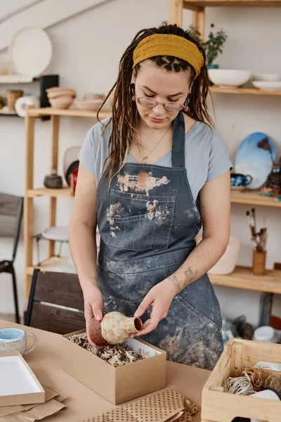 職人の若い女性は 手作りの粘土花瓶を取るか 工芸品のためのさまざまな供給とテーブルのそばに立っている間 箱からジャグ — ストック写真