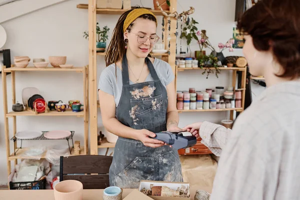 小さな陶器店の若い女性の所有者クレジットカードで購入のために支払う顧客に手作りのアイテムを販売する決済端末を持つ — ストック写真