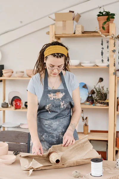 陶瓷店的年轻女工站在桌旁用包装材料把手工制作的陶罐或花瓶包在纸上 — 图库照片