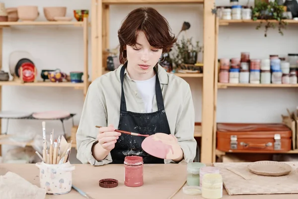 作業着の若い創造的な女性供給品と棚に対してテーブルに座っている間マルサラ色の手作りの陶器を塗装 — ストック写真