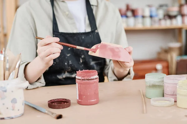 テーブルの上にペインブラシと粘土ボウルを保持エプロンの若い創造的な女性は マルサラで手作りの陶器を着色しながら — ストック写真