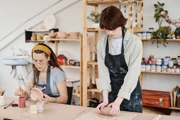若い女性ですエプロン混練作品の柔らかい粘土のテーブルによって彼女の同僚に対して絵筆や陶器の着色ボウルと — ストック写真
