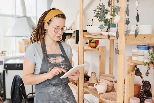 新しい粘土アイテムと棚の前に立っている間 顧客のオンライン注文を見て土器店の若い女性の所有者 — ストック写真