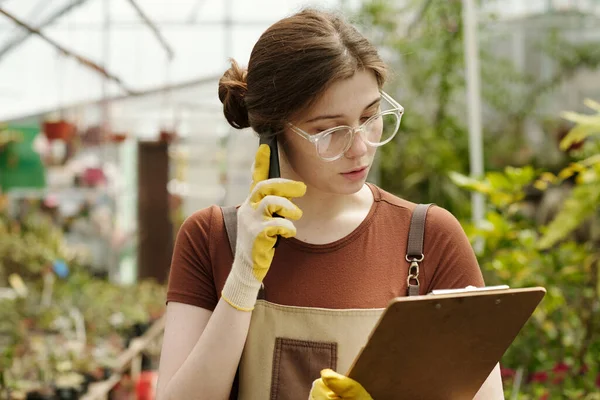 携帯電話で顧客と話をしながら 文書によると花の可用性を確認眼鏡の若い女性花屋 — ストック写真