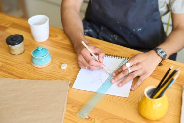 Önlüklü genç adamın elleri ahşap masada oturuyor ve çizim yapıyor. Kalemle ve not defterinin sayfasında yaratıcı kilden bir kalem ve cetvelle.