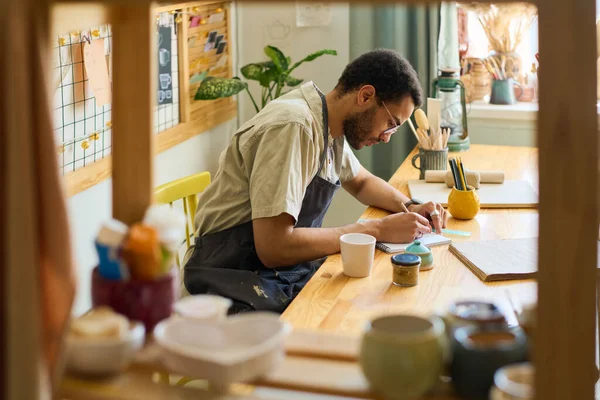 Young creative man bending over workplace while making sketch in notepad in spacious studio or workshop with displays for new items