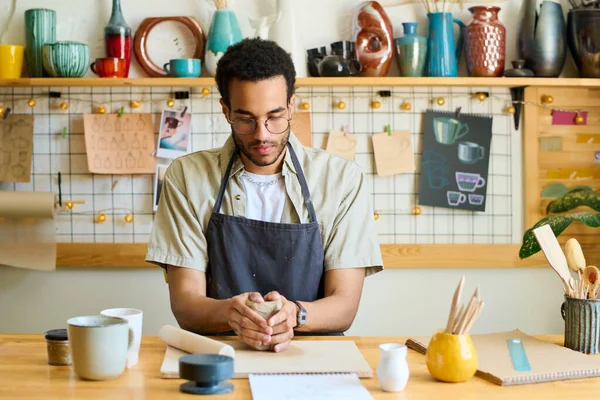 ワークショップやスタジオでテーブルに座り ボード上の新しい粘土アイテムを作成することに熟練した若い創造的な黒人男性 — ストック写真