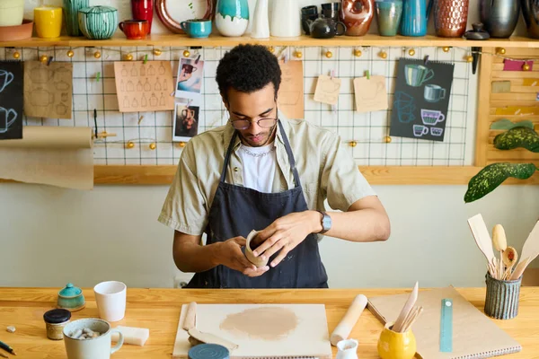 作業着を着た若いクリエイティブ ブラックマンが 作業場の陶器の棚にテーブルに座りながら販売する新しい粘土アイテムを作成します — ストック写真