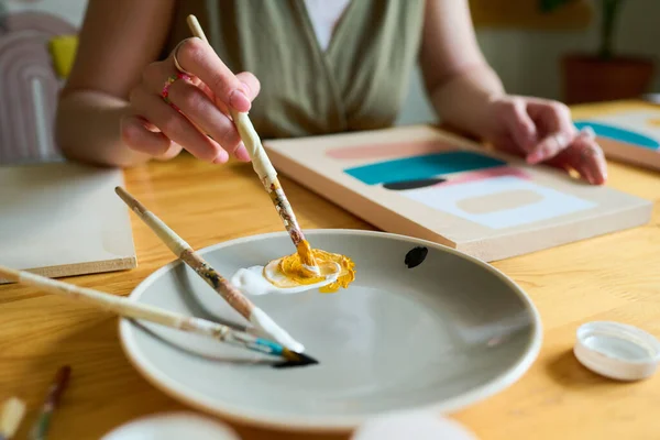 テーブルに座り 紙に絵を描く一方で プレート上に白と黄色を混合筆で若い創造的な女性の手 — ストック写真