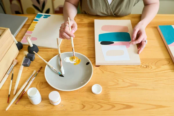 木製のテーブルに座って紙の上に抽象的な絵を描きながら プレート上に色を混合若い女性アーティストの手の角度の上に — ストック写真