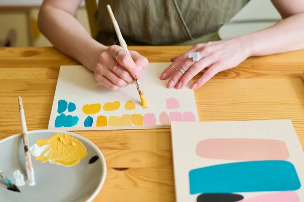 ワークショップで木製のテーブルに座っている間 彼女の新しい絵のための色の腕時計を作る絵筆を持つ若い女性の手 — ストック写真