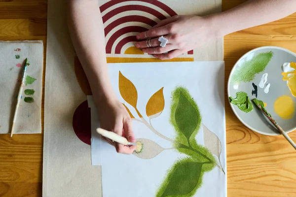 ワークショップでテーブルに座りながら 筆で緑の葉を紙に描く若い女性アーティストの手の角度の上に — ストック写真