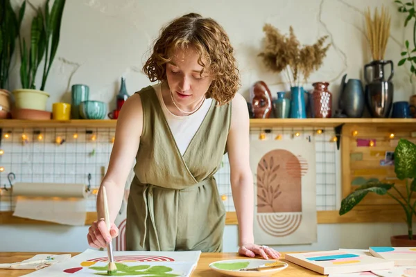 若い創造的な女性がテーブルの上に曲げながら ワークショップで絵筆と緑の水彩で紙に抽象的な絵を描く — ストック写真