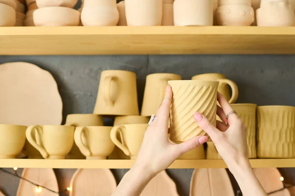 店内の他の土器と棚の上の品揃えから1つを選択しながら 手作りの粘土マグカップを保持若い女性の手 — ストック写真