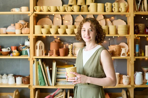 Young successful owner of small shop with handmade clay mug in hands standing by display with earthenware created for sale