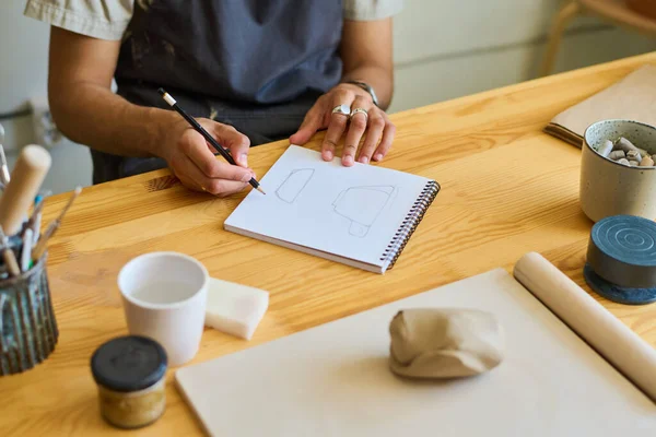 粘土製品を作るための消耗品とテーブルに座っている間ノートパッドのページにカップのスケッチを作る若い創造的な職人の手 — ストック写真