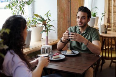 Elinde kahve olan bir adam ve kız arkadaşı masanın önünde oturmuş tatlı tatlı kahve içerken sohbet ediyorlar.