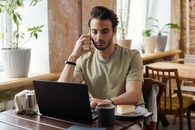 Kafe 'de dizüstü bilgisayarın önünde oturup sohbet ederken ve kahve içerken günlük kıyafetlerle serbest çalışan genç bir erkek.