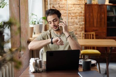 Kafe 'de dizüstü bilgisayarın önünde oturup, kol saatine ve ağ kurma sistemine bakarken, günlük kıyafetlerle meşgul genç bir adam cep telefonuyla konuşuyor.