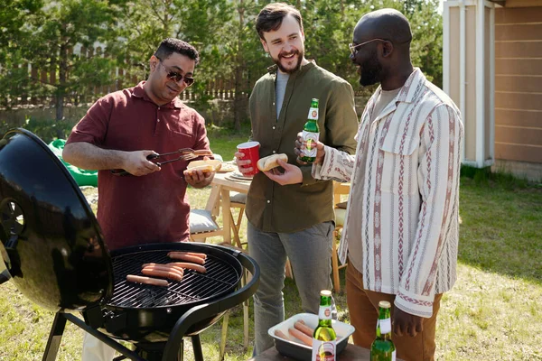 在周末的户外派对上 三位快乐的跨文化年轻人一边喝啤酒一边聊天 其中一位正在烧烤 — 图库照片