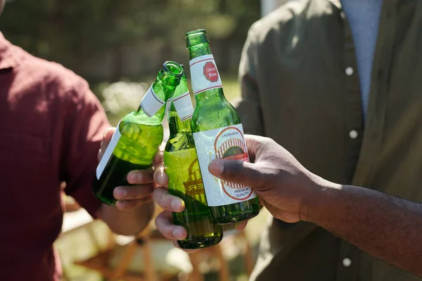 stock image Hands of three intercultural men clinking with bottles of beer during outdoor party on weekend and enjoying their meeting