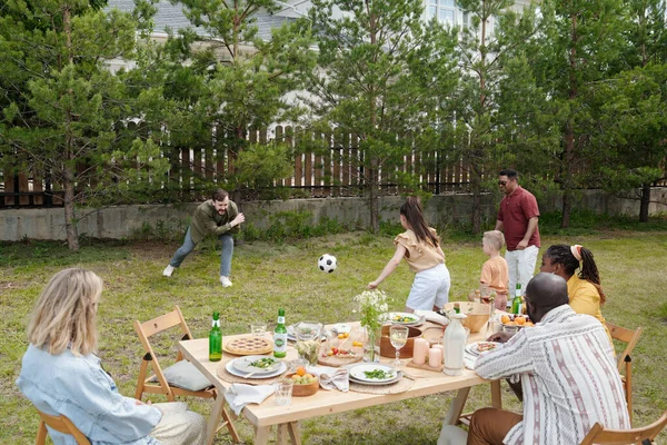 年轻的黑人夫妇和白人妇女坐在餐桌旁 看着两个不同文化的男人和孩子在户外玩游戏 — 图库照片