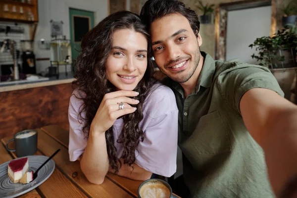 在浪漫的约会中 这对快乐的年轻夫妇一边在咖啡馆里吃着芝士蛋糕和卡布奇诺 一边看着笑脸的男人拿着智能手机相机 — 图库照片