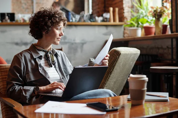 深刻なブルネットビジネスマン女性またはフリーランサーともに紙で手とラップトップ上の彼女の膝の上に座ってテーブルでカフェと分析財務データ — ストック写真