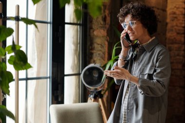 Genç bayan yönetici ya da günlük giyimli kafe sahibi büyük pencerenin önünde dururken müşterilerle cep telefonuyla konuşuyor.