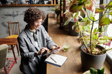 Tabletli kafenin genç bayan sahibi pencerenin yanında otururken web sitesinde veya yeni müşteri siparişleri arasında geziniyor