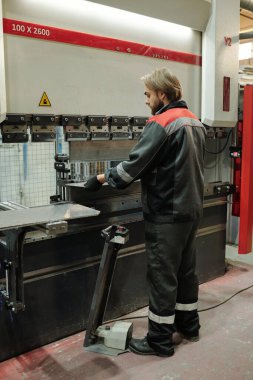 Atölyede büyük bir endüstriyel makinenin önünde çalışırken iş kıyafetleri içindeki genç bir adamın mekanizma pedalını iterken yan görünümü