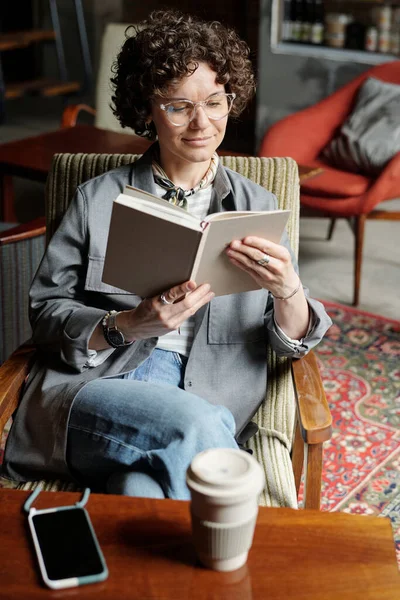 居心地の良いカフェで快適なアームチェアに座っている間 自分の前で開いて本を保持暗い巻き毛を持つ若い安らかな女性 — ストック写真