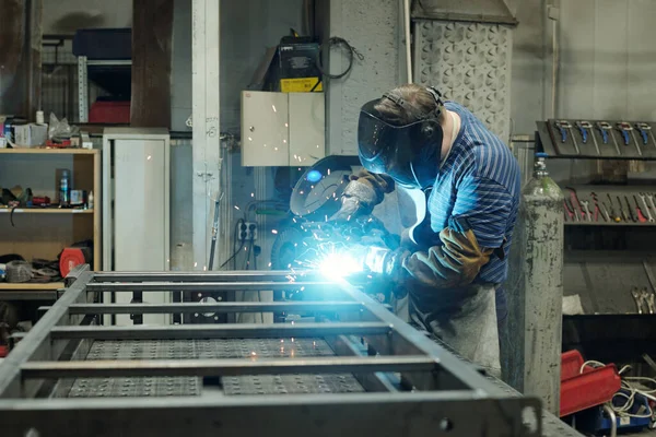 金属作業台の上に曲げながら修理や建設作業を行う保護面の若い男性溶接機の側面図 — ストック写真