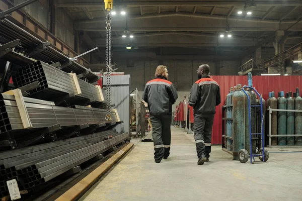 倉庫内の通路に沿って移動しながら 新しい機器の品質を議論制服を着た2人の若い異人種間の男性労働者の背面図 — ストック写真