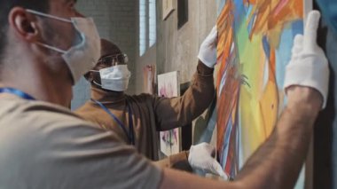 Beyaz eldivenli ve yüz maskeli iki gencin sanat galerisindeki duvarda soyut sanat eserleri asılı duran ve konuşan iki genç adamın yan görüntüsü.