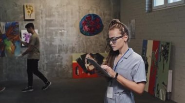Sanat galerisinde duran, tablet bilgisayar kullanan, kameraya bakıp gülümseyen genç beyaz bir kadının belden yukarı portresi. Duvara soyut bir resim asmış iki adam.