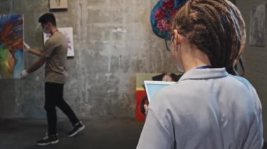 Sanat galerisinde ayakta duran, tablet bilgisayar kullanan, maskeli ve eldivenli, duvarında soyut bir resim olan iki erkek, genç bayan yöneticinin omuzları üzerinde.
