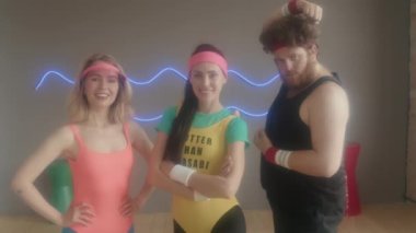 Koyu saçlı beyaz kadın eğitmenin portresi, 80 'lerin aerobik sınıfındaki genç adam ve kadın gündüz kameraya bakıyor ve poz veriyor.