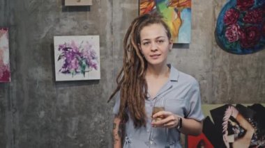 Elinde bir kadeh şampanya tutan, modern sanat galerisinde durup kameraya gülümseyen neşeli beyaz kızın orta boy portresi.