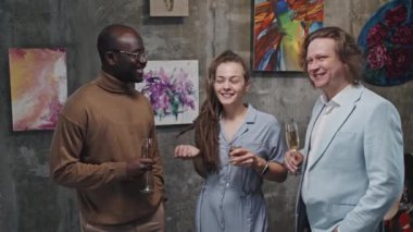 Rastalı neşeli beyaz bir kızın orta boy portresi. Elinde şampanya kadehleri olan iş adamı ve siyah bir adam. Modern sanat galerisinde duruyor, kameraya bakıyor ve gülümsüyor.